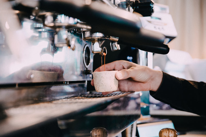 Image of a barista monitoring the extraction of Espresso shots on a La Marzocco Linea Classic coffee machine. The Barista also uses tools such as coffee tampers, tamping mats, milk steaming pitchers, milk pitcher rinsers and knock out drawers.